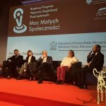 Panel dyskusyjny podczas konferencji Programu Moc Małych Społeczności. Na scenie siedzi kilka osób uczestniczących w dyskusji, jedna z nich trzyma mikrofon. W tle widoczny ekran z nazwą programu oraz logotypami instytucji, a po bokach stoją udekorowane choinki.