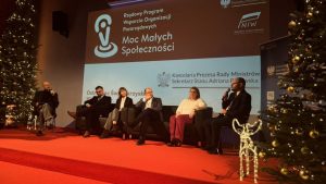 Panel dyskusyjny podczas konferencji Programu Moc Małych Społeczności. Na scenie siedzi kilka osób uczestniczących w dyskusji, jedna z nich trzyma mikrofon. W tle widoczny ekran z nazwą programu oraz logotypami instytucji, a po bokach stoją udekorowane choinki.