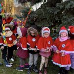Grupa dzieci ubranych w czerwone stroje i czapki świąteczne stoi przy choince na świeżym powietrzu. Dzieci trzymają papierowy łańcuch i drobne ozdoby choinkowe, a dorosła osoba pomaga im w dekorowaniu drzewa.