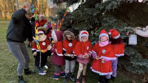 Grupa dzieci ubranych w czerwone stroje i czapki świąteczne stoi przy choince na świeżym powietrzu. Dzieci trzymają papierowy łańcuch i drobne ozdoby choinkowe, a dorosła osoba pomaga im w dekorowaniu drzewa.