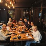 Grupa seniorów siedzi przy wspólnym stole w restauracji podczas spotkania integracyjnego. Uczestnicy rozmawiają, jedzą posiłki i spędzają razem czas w atmosferze refleksji i podziękowań dla członków Klubów Seniora oraz wolontariuszy.