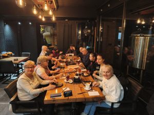 Grupa seniorów siedzi przy wspólnym stole w restauracji podczas spotkania integracyjnego. Uczestnicy rozmawiają, jedzą posiłki i spędzają razem czas w atmosferze refleksji i podziękowań dla członków Klubów Seniora oraz wolontariuszy.