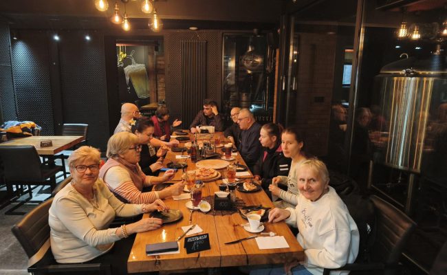 Grupa seniorów siedzi przy wspólnym stole w restauracji podczas spotkania integracyjnego. Uczestnicy rozmawiają, jedzą posiłki i spędzają razem czas w atmosferze refleksji i podziękowań dla członków Klubów Seniora oraz wolontariuszy.