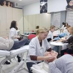 Zajęcia praktyczne w sali szkoleniowej. Kilka osób w fartuchach wykonuje zabiegi pielęgnacyjne na twarzy i dłoniach u modeli leżących lub siedzących na stanowiskach. W tle widoczne są szafki, lustra oraz sprzęt wykorzystywany podczas zajęć.