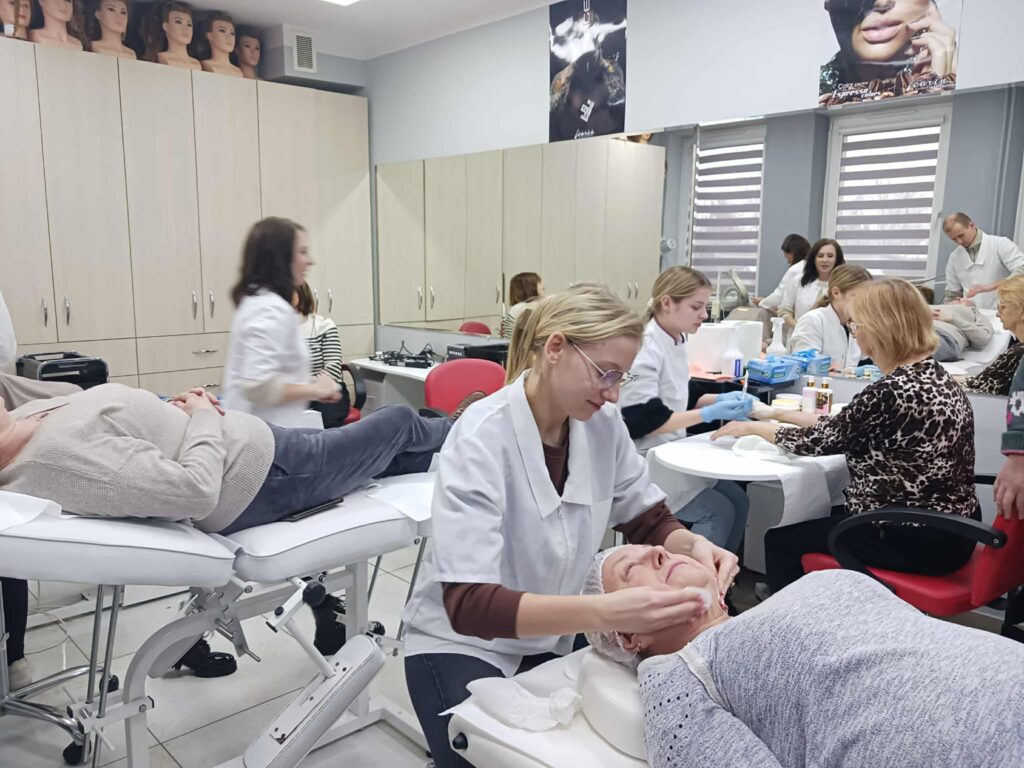 Zajęcia praktyczne w sali szkoleniowej. Kilka osób w fartuchach wykonuje zabiegi pielęgnacyjne na twarzy i dłoniach u modeli leżących lub siedzących na stanowiskach. W tle widoczne są szafki, lustra oraz sprzęt wykorzystywany podczas zajęć.